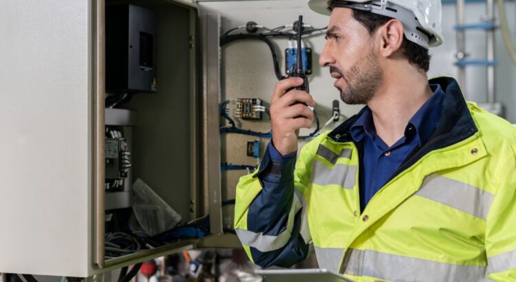 Mecânico de manutenção industrial analisando painel elétrico com rádio comunicador e tablet, realizando inspeção técnica e monitoramento de equipamentos industriais.